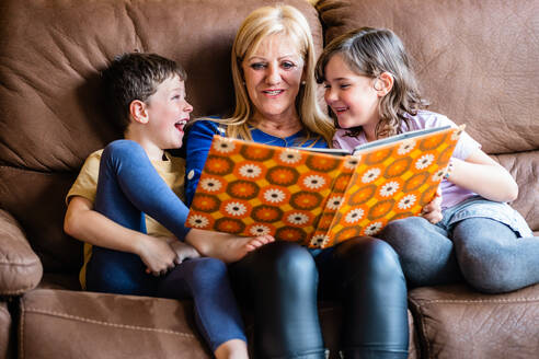 Glückliche reife Großmutter, die lächelt und ein interessantes Buch für liebenswerte Kinder liest, während sie zusammen auf einer bequemen Couch sitzen - ADSF44002