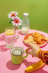 Unbekannte Person trinkt heißen aromatischen Kaffee in einer Tasse, die auf einem Tisch mit leckeren Croissants und Bananen steht, der mit Blumen in einer Vase dekoriert ist - ADSF44046