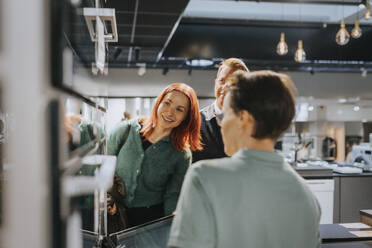 Happy couple looking at microwave oven while saleswoman assisting at electronics store - MASF36675