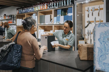 Female sales clerk giving appliance box to customer at checkout counter in electronics store - MASF36683