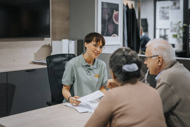 Smiling female interior designer explaining senior couple over brochure at desk in store - MASF36696