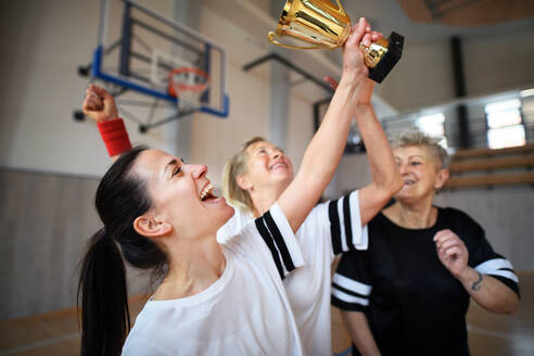 Eine Gruppe junger und alter Frauen, Basketballspielerinnen, in der Turnhalle mit einem Pokal, die den Sieg feiern. - HPIF09379