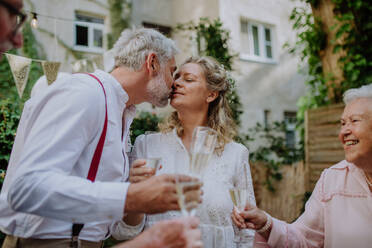 Reife Braut und Bräutigam küssen sich bei einem Hochzeitsempfang mit ihrer Familie, draußen im Garten. - HPIF09555