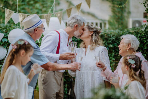 Reife Braut und Bräutigam küssen sich bei einem Hochzeitsempfang mit ihrer Familie, draußen im Garten. - HPIF09558