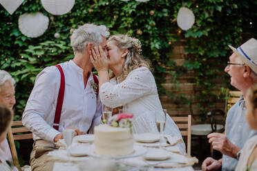 Reife Braut und Bräutigam küssen sich bei einem Hochzeitsempfang mit ihrer Familie, draußen im Garten. - HPIF09570