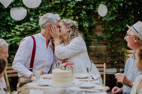 Reife Braut und Bräutigam küssen sich bei einem Hochzeitsempfang mit ihrer Familie, draußen im Garten. - HPIF09570