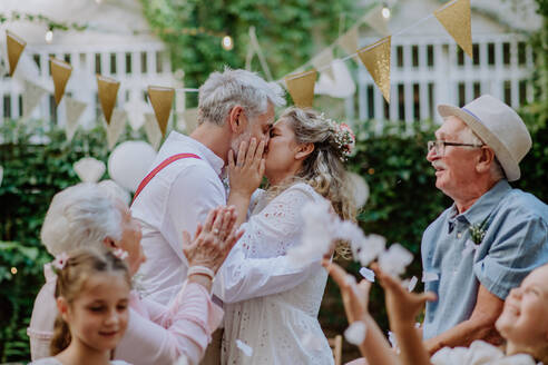 Reife Braut und Bräutigam küssen sich bei einem Hochzeitsempfang mit ihrer Familie, draußen im Garten. - HPIF09572