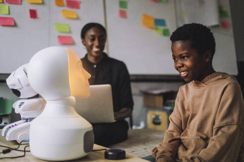 Glückliche Studentin, die mit einem beleuchteten sozialen Roboter im Innovationslabor kommuniziert - MASF36748