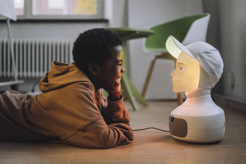 Fröhlicher Junge vor einem beleuchteten KI-Roboter, der im Innovationslabor auf dem Boden liegt - MASF36756