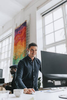 Lächelnder junger Geschäftsmann, der einen Desktop-PC am Schreibtisch im Büro benutzt - MASF36785