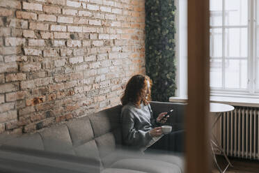 Geschäftsfrau, die während der Kaffeepause im Büro auf dem Sofa sitzend ihr Smartphone benutzt - MASF36826