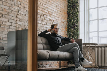 Businessman talking on mobile phone while sitting on sofa in office - MASF36828