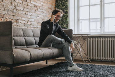 Smiling businessman talking on smart phone while sitting on sofa at office - MASF36829