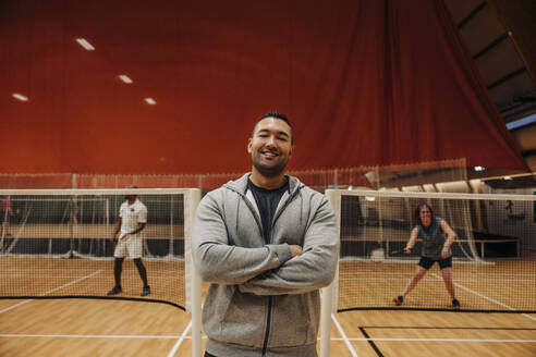 Lächelnder männlicher Trainer, der mit verschränkten Armen auf dem Badmintonplatz steht - MASF36846