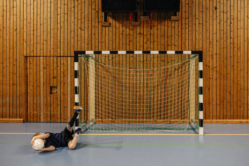 Male goalie with disabled leg lying on floor holding football while playing at sports court - MASF36866