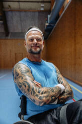 Porträt eines selbstbewussten männlichen Sportlers, der mit verschränkten Armen auf einem Sportplatz im Rollstuhl sitzt - MASF36878