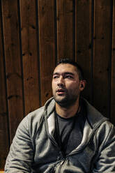 Male athlete looking away while sitting near wooden wall - MASF36882