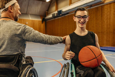 Male athlete with disability motivating girl sitting on wheelchair with basketball at sports court - MASF36893