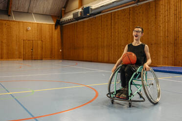 Happy girl with basketball sitting on wheelchair at sports court - MASF36894