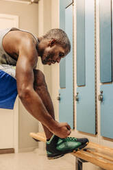 Male athlete tying shoe lace by bench in locker room - MASF36910