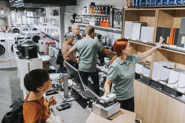 Female sales clerk showing various appliances on shelf to customer at checkout counter - MASF36923
