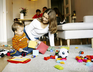 Lächelnde Mutter sitzt neben ihrem Sohn, der zu Hause mit Spielzeug spielt - JJF00959