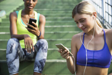 Frau in Sportkleidung benutzt Smartphone mit Freund auf Stufen sitzend - IKF00449