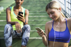 Frau in Sportkleidung benutzt Smartphone mit Freund auf Stufen sitzend - IKF00449