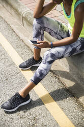 Woman wearing sports shoes using smart phone sitting by road - IKF00485