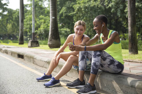 Glückliche Freunde, die sich ein Smartphone teilen, sitzen auf dem Gehweg im Park - IKF00486