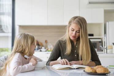 Mutter liest ein Buch und sitzt mit ihrer Tochter in der Küche zu Hause - IKF00494