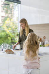 Mädchen lehnt auf Kücheninsel mit Mutter bereitet Pizza im Hintergrund - IKF00496