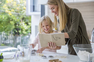 Glückliche Frau, die ein Rezeptbuch liest, während ihre Tochter in der Küche zu Hause Essen zubereitet - IKF00498