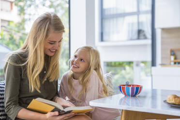 Lächelndes Mädchen, das seine Mutter beim Lesen eines Buches in der Küche zu Hause betrachtet - IKF00499