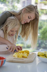 Smiling woman assisting daughter cutting pineapple on cutting board at home - IKF00519