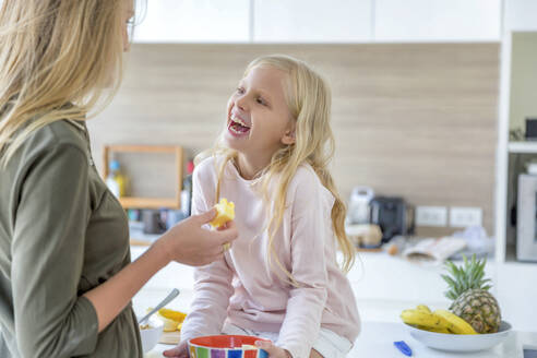 Glückliche Tochter schaut Mutter an, die zu Hause eine Ananas hält - IKF00521