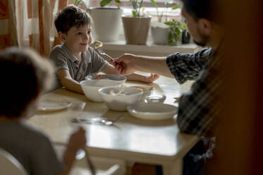 Father feeding dumpling to son at dining table in home - ANAF01395