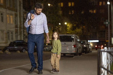 Vater hält die Hand seines Sohnes, der nachts auf dem Bürgersteig in der Stadt aufwacht - ANAF01396