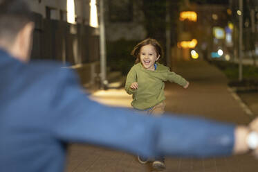 Cheerful son running towards father on sidewalk at night - ANAF01400