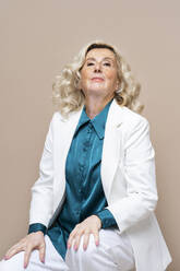 Confident senior businesswoman sitting against beige background - AAZF00441