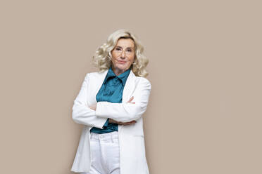 Smiling blond senior businesswoman with arms crossed against beige background - AAZF00443