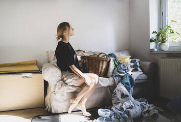 Nachdenkliche Frau auf dem Sofa sitzend mit unordentlicher Wäsche zu Hause - NDEF00612