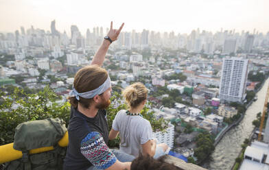 Man gesturing horn sign with woman looking at city view from rooftop - IKF00580