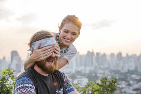 Playful woman covering man eyes with city in background - IKF00582