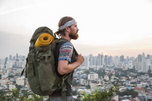 Junger Mann mit Rucksack vor einer Stadtlandschaft bei Sonnenuntergang - IKF00599