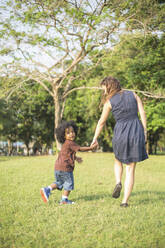 Mother and son holding hands and walking on grass at park - IKF00618