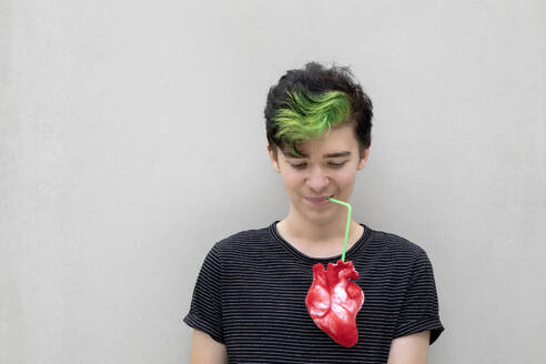 Jugendlicher mit grün gefärbten Haaren, der aus einem Herz trinkt, vor grauem Hintergrund - PSTF01068
