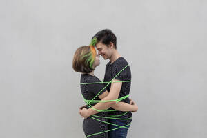 Teenage couple tied up with green rope embracing each other against gray background - PSTF01072