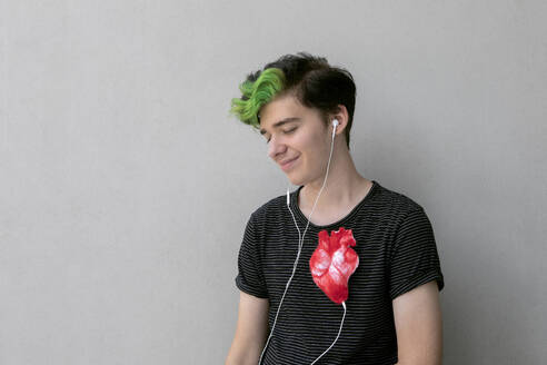Teenage boy listening heartbeat with headphones connected to heart against gray background - PSTF01079