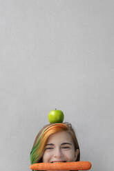 Teenager-Mädchen balanciert Apfel auf dem Kopf mit Karotte im Mund vor grauem Hintergrund - PSTF01082